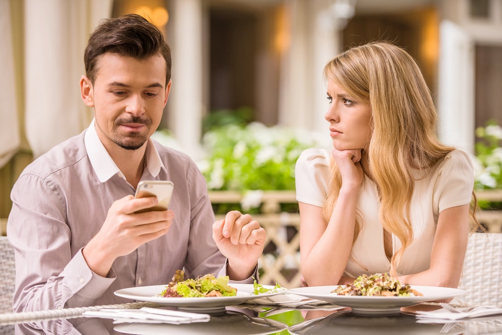 This is a photo of a couple at a restaurant. The man is on his phone, and the woman with him looks unhappily over to him