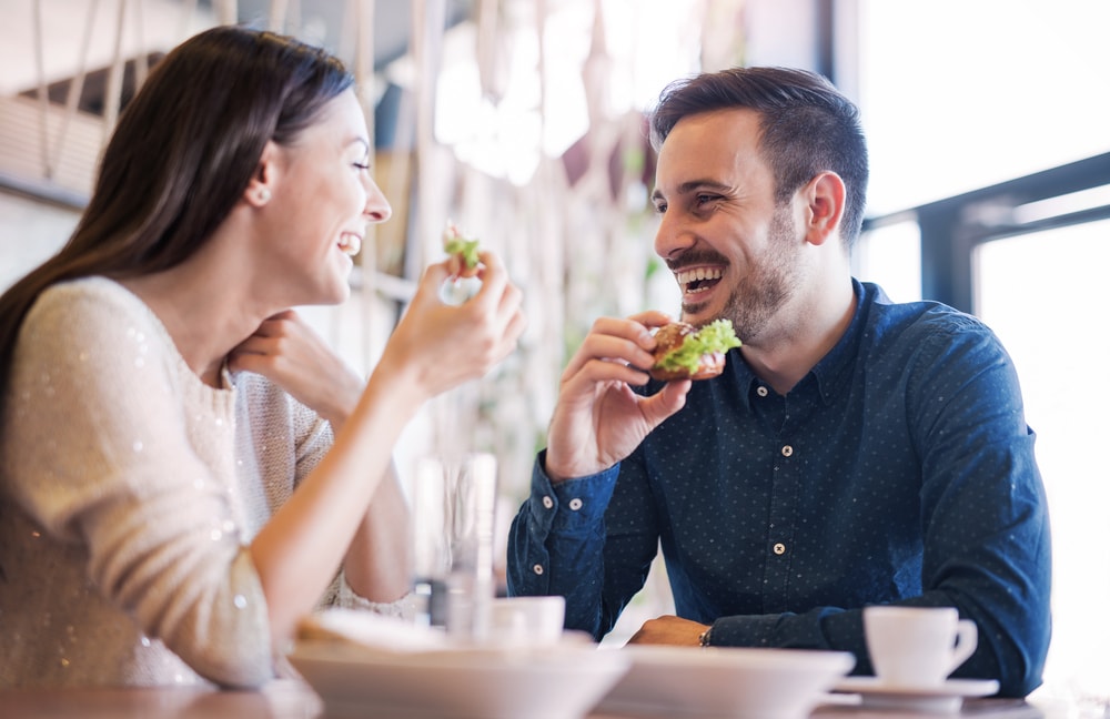 This is a photo of a happy couple at a restaurant. They are both laughing mid-conversation, holding sandwiches