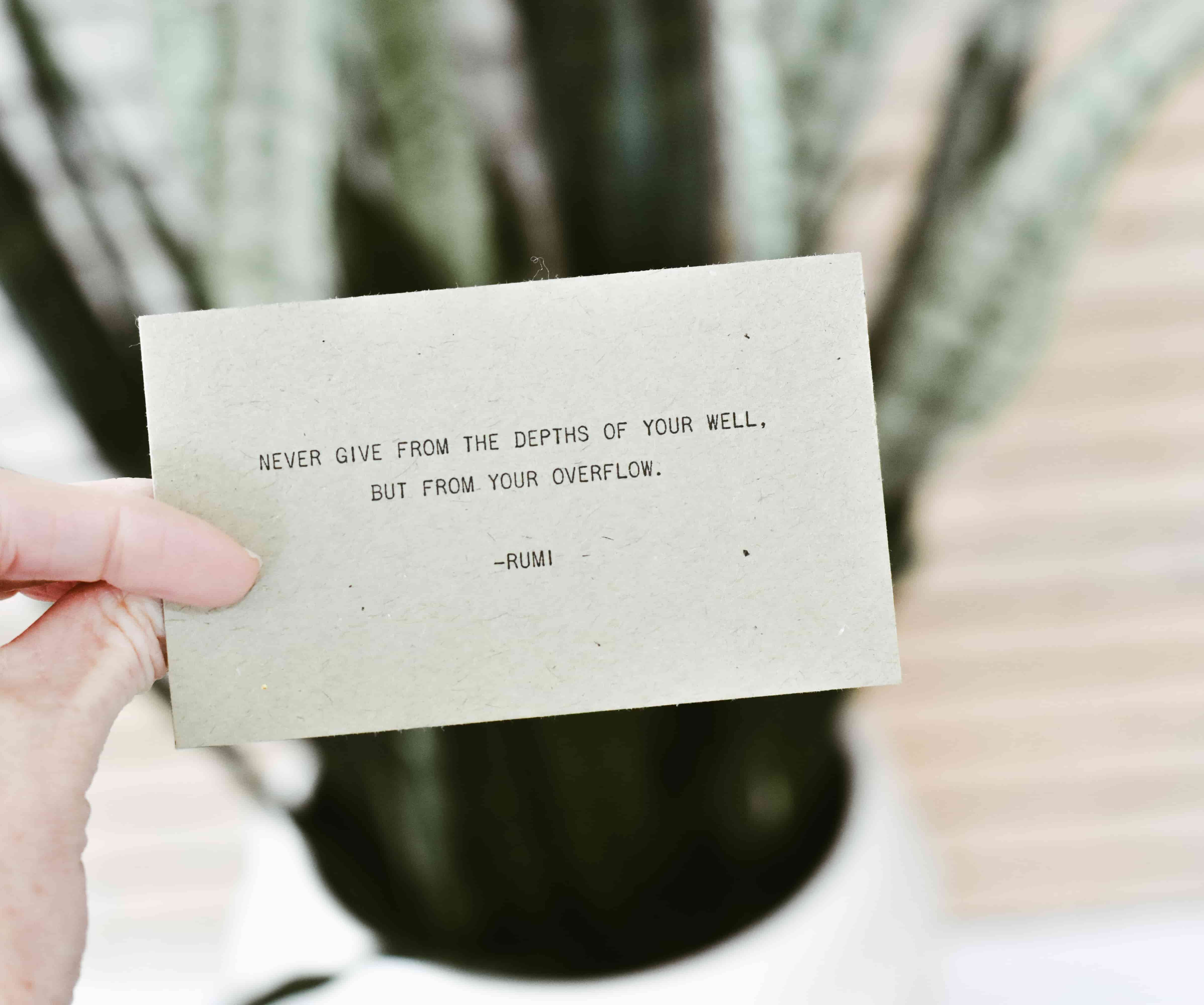 A hand holds an affirmation card with a quote by Rumi. It says 'never give from the depths of your well, but from your overflow'.