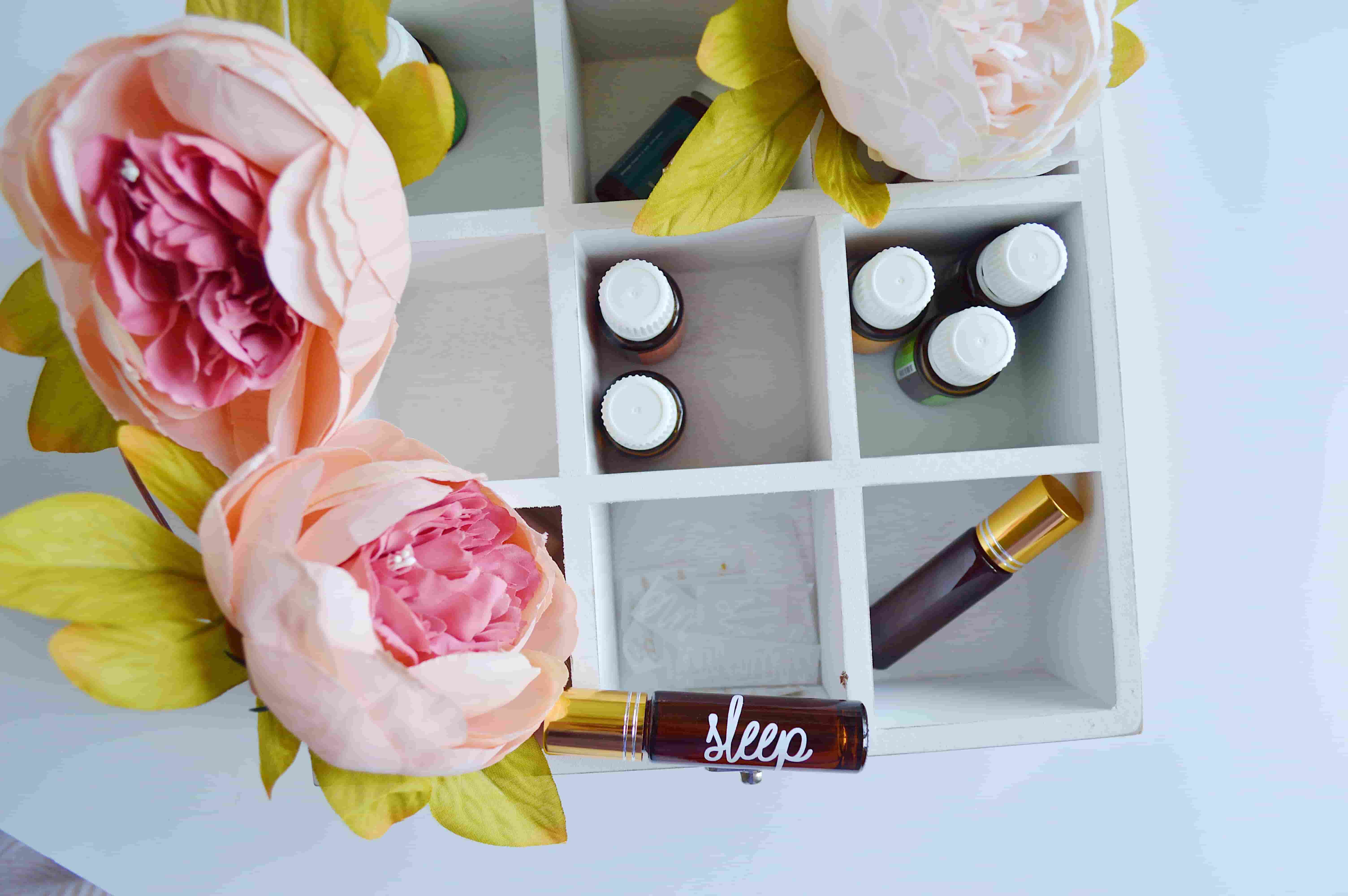 A white painted box has natural pink blossoms resting against it. Within the box, aromatherapy oils labelled with scents and the word 'sleep' are resting.