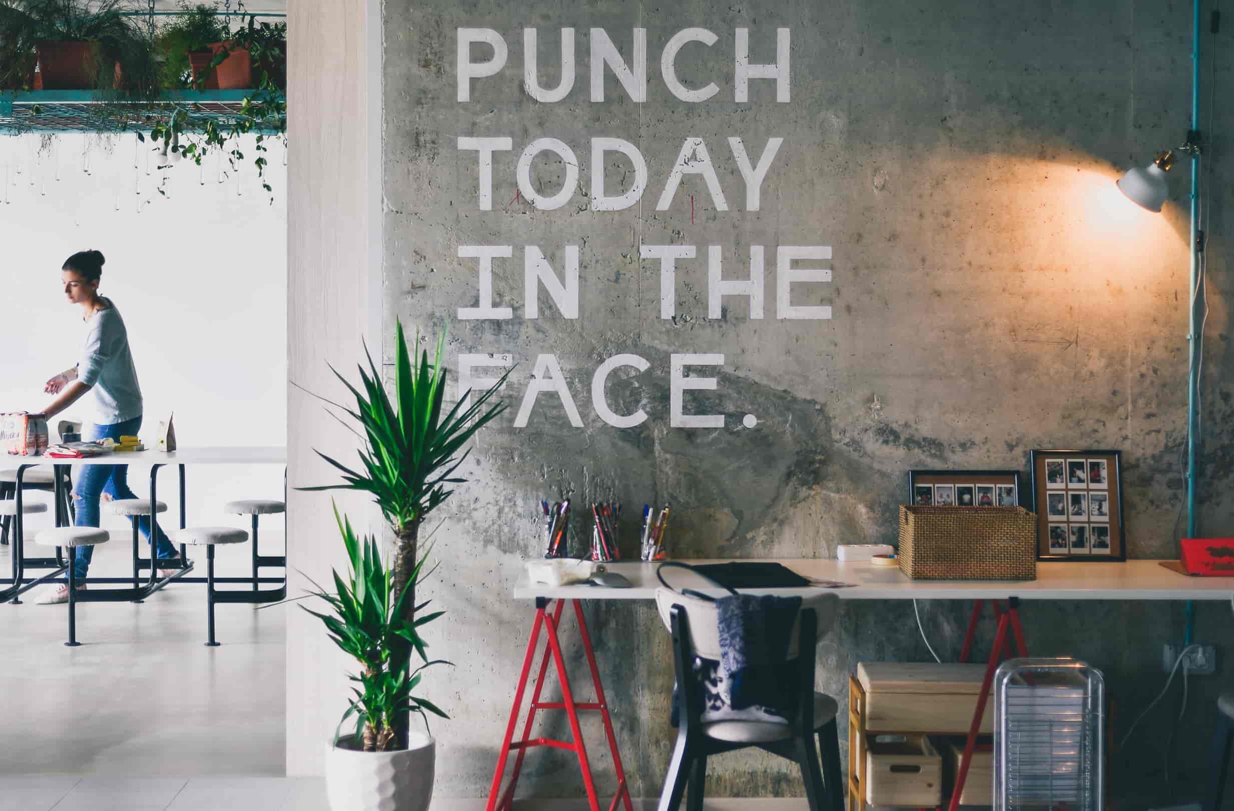 A motivational quote is written on a bare concrete wall in a modern office. It says: punch today in the face.