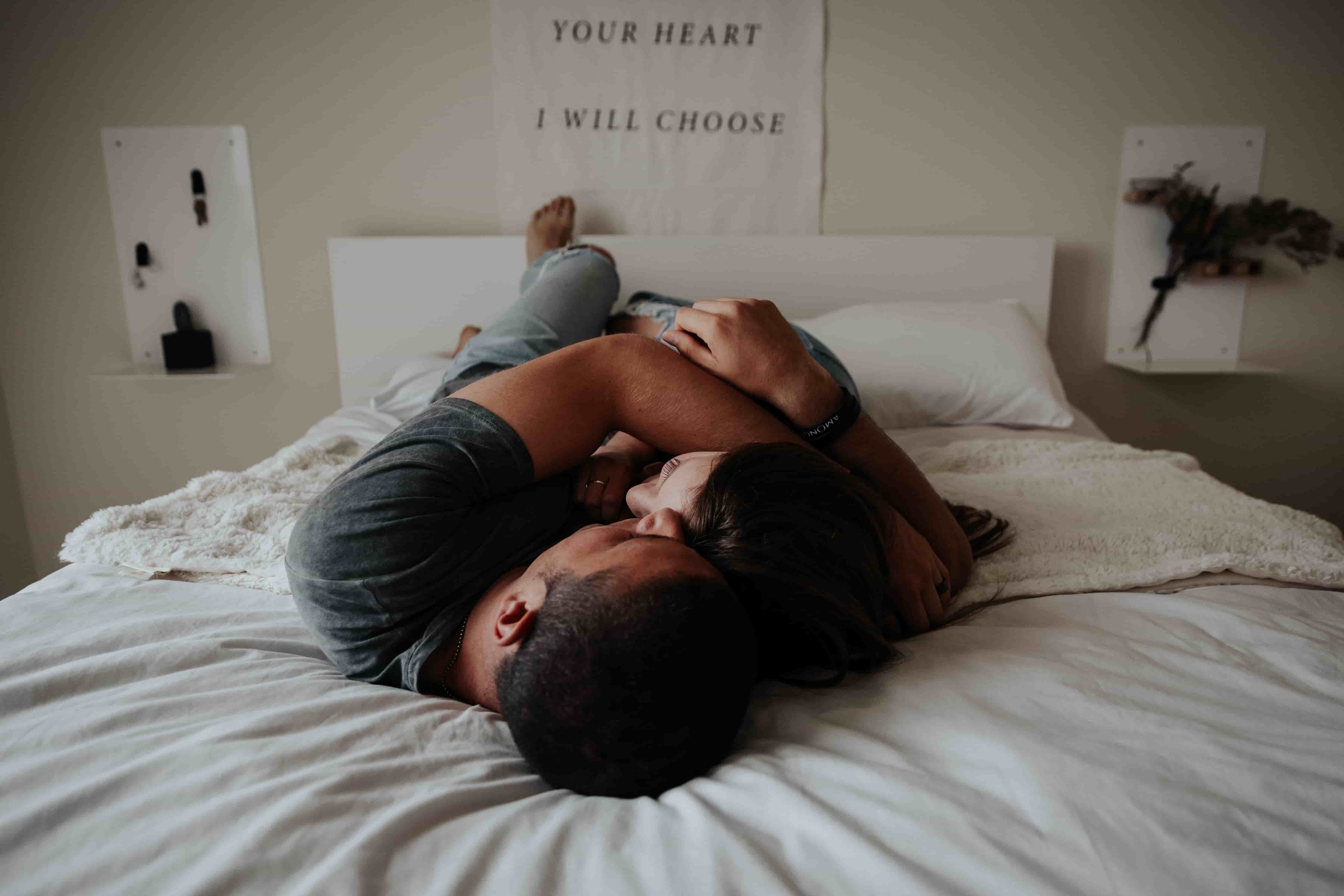 A couple lay together in bed, holding each other close and looking sad.
