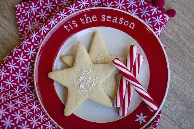 A close-up of peppermint sticks and star-shaped cookies on a plate that says 'tis the season'