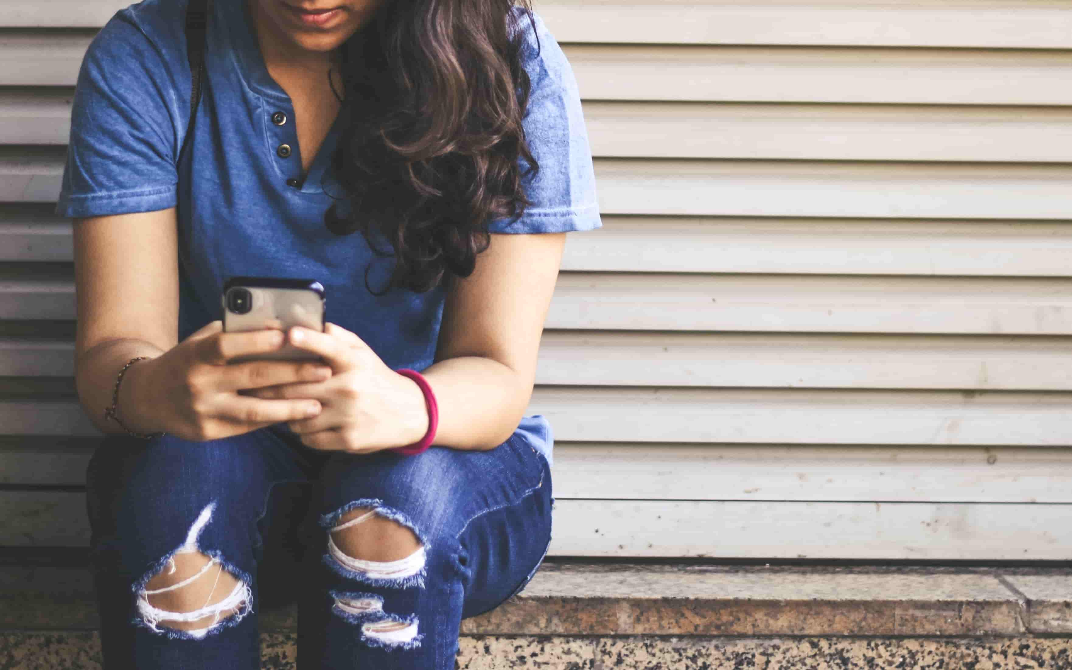 A woman sits outside on a step, holding her phone in both hands as she texts with a nervous look.