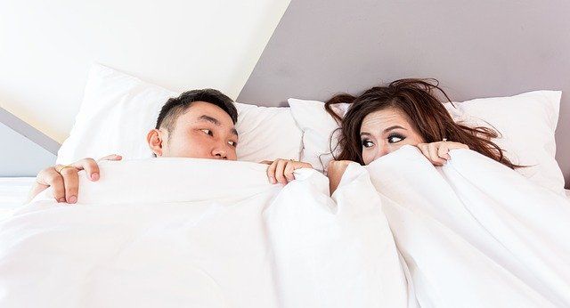 A couple look at each other nervously, peeking out from under a duvet