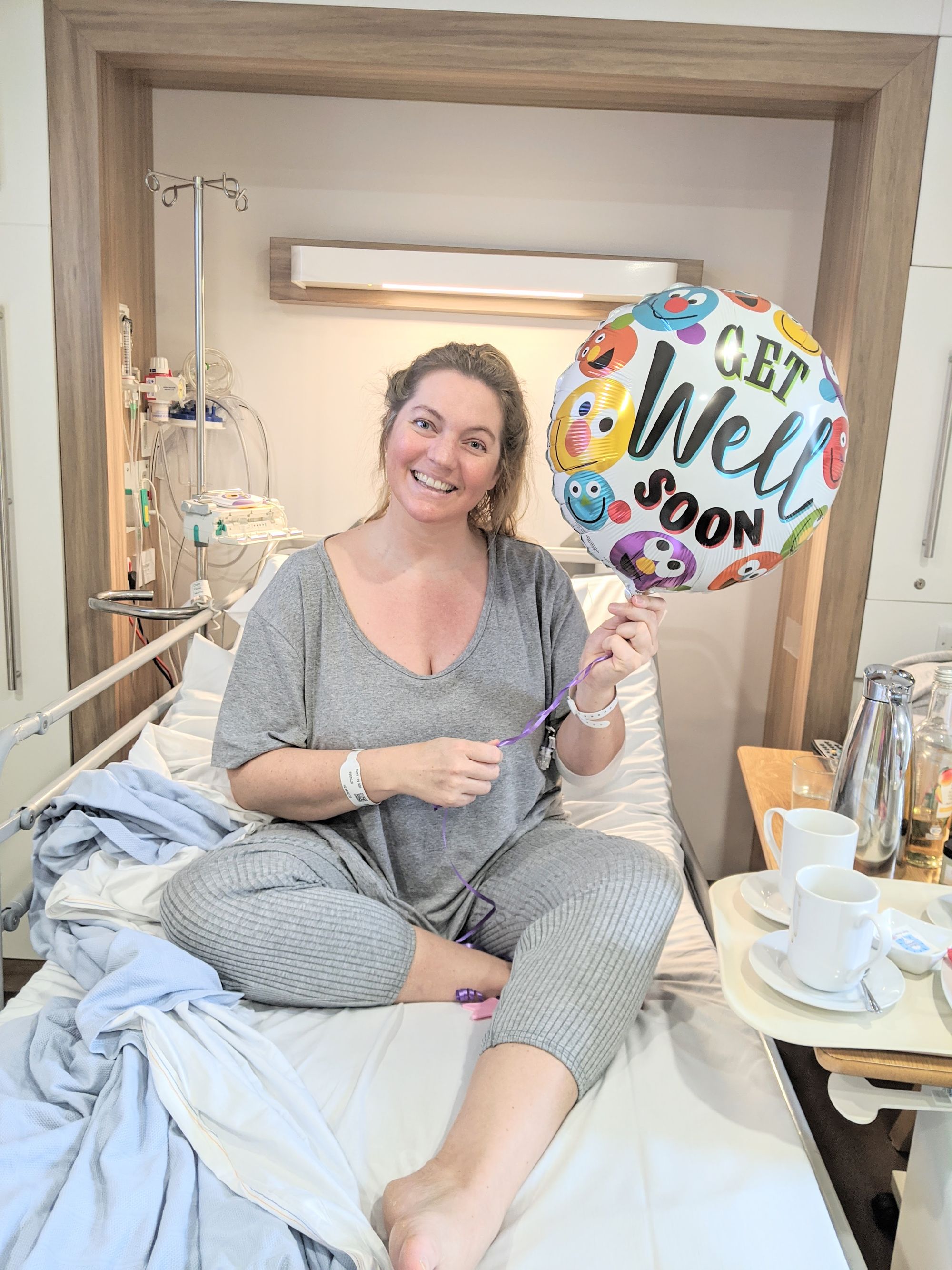 Beth in hospital smiling and holding a "Get well soon" balloon