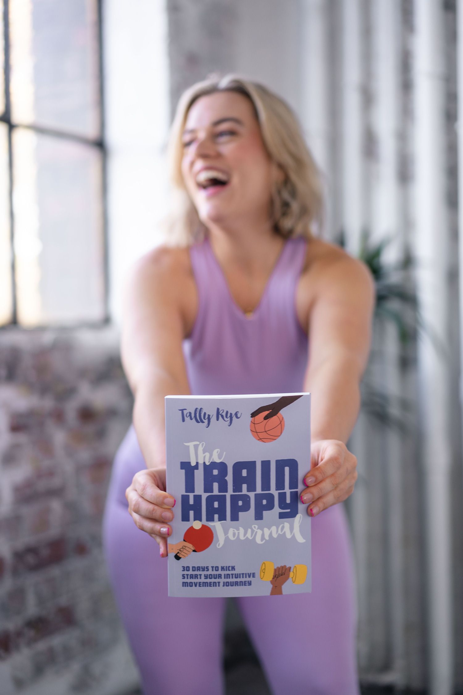 Tally Rye is in the background smiling, she is holding out her new book The Train Happy Journal