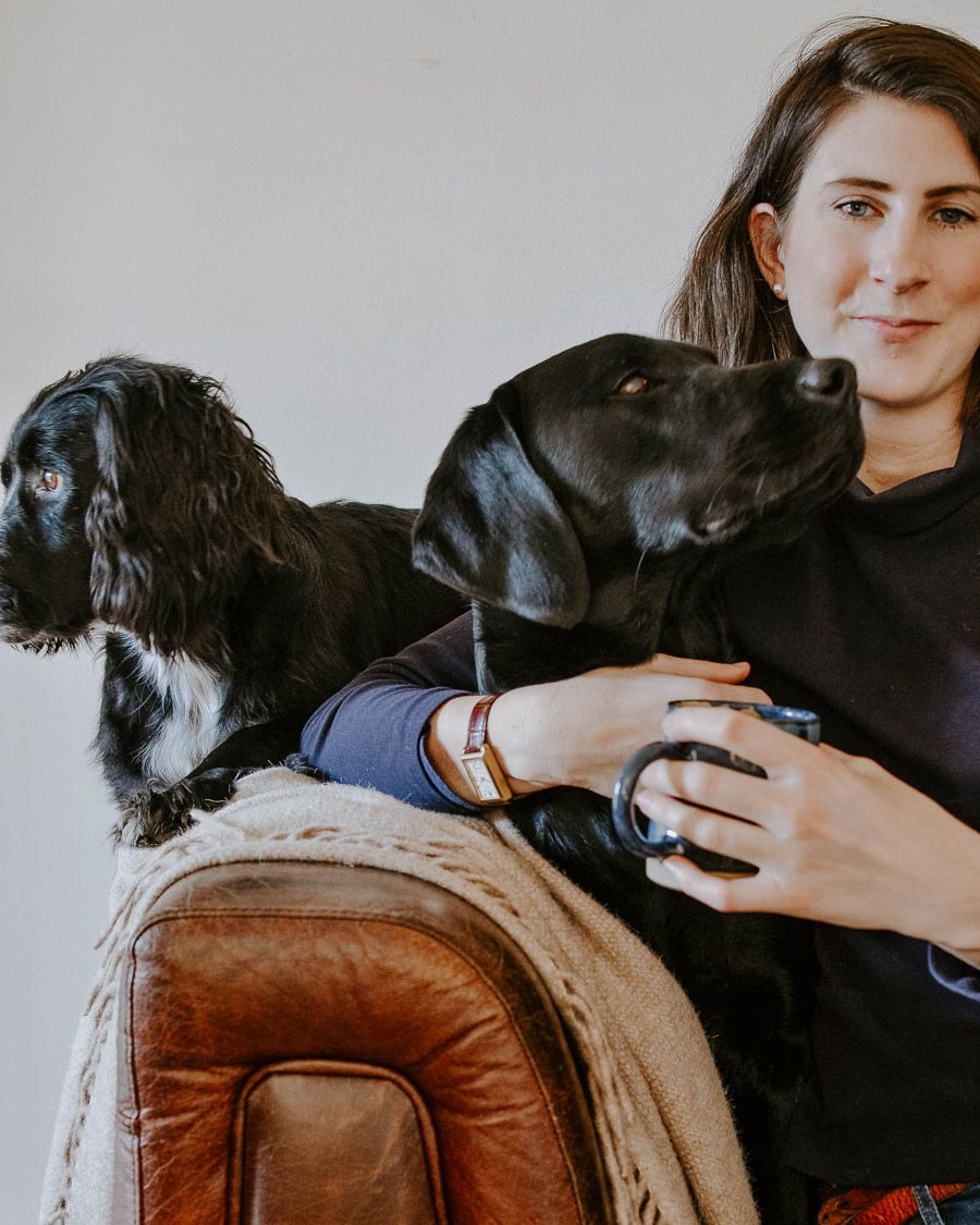 Josephine sitting on a sofa with a mug of tea and her two black dogs are sat on her lap. Josephine is slightly smiling at the camera